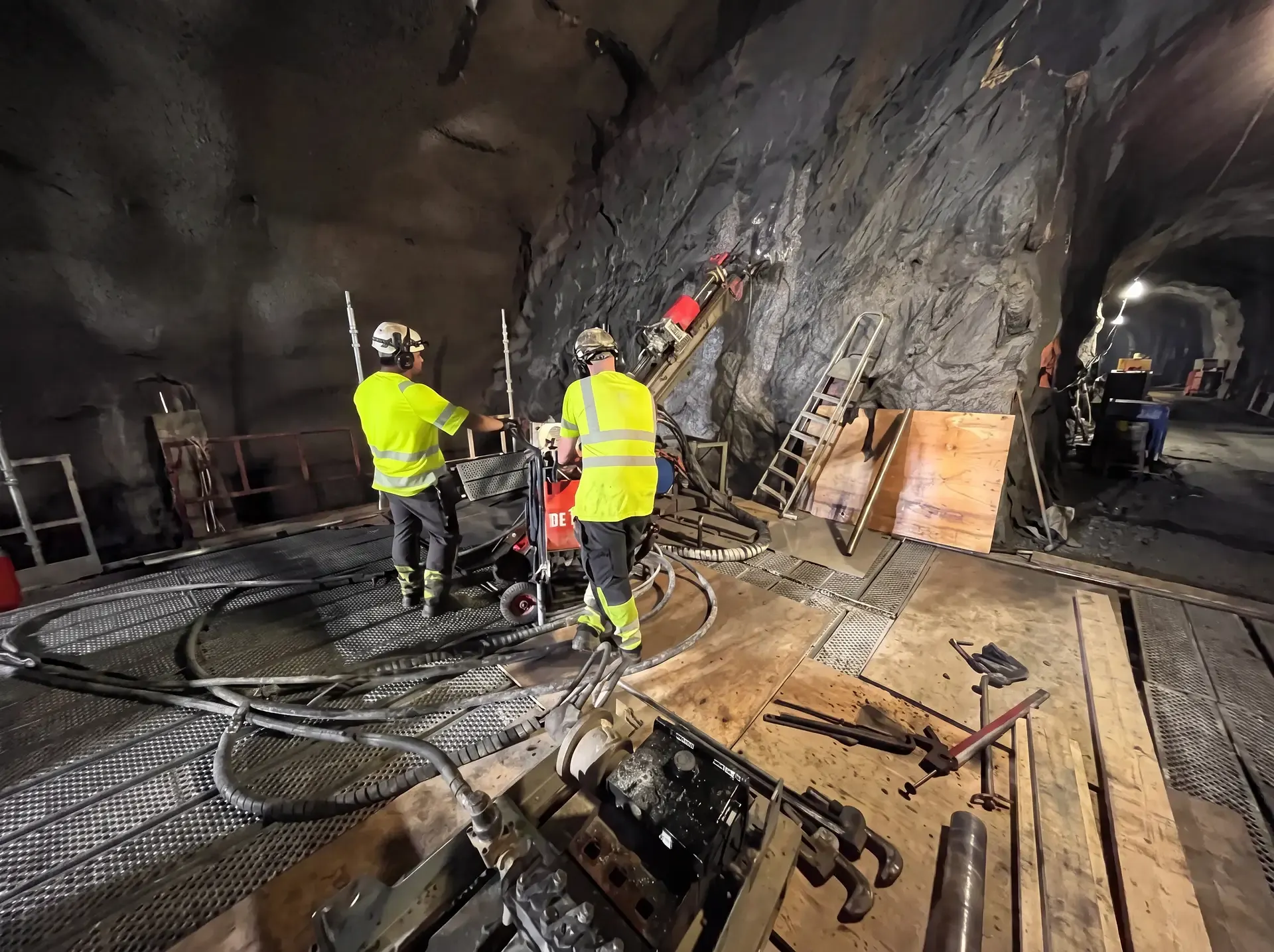 Två gruvarbetare i varselkläder arbetar med borrutrustning i bergtunnel.