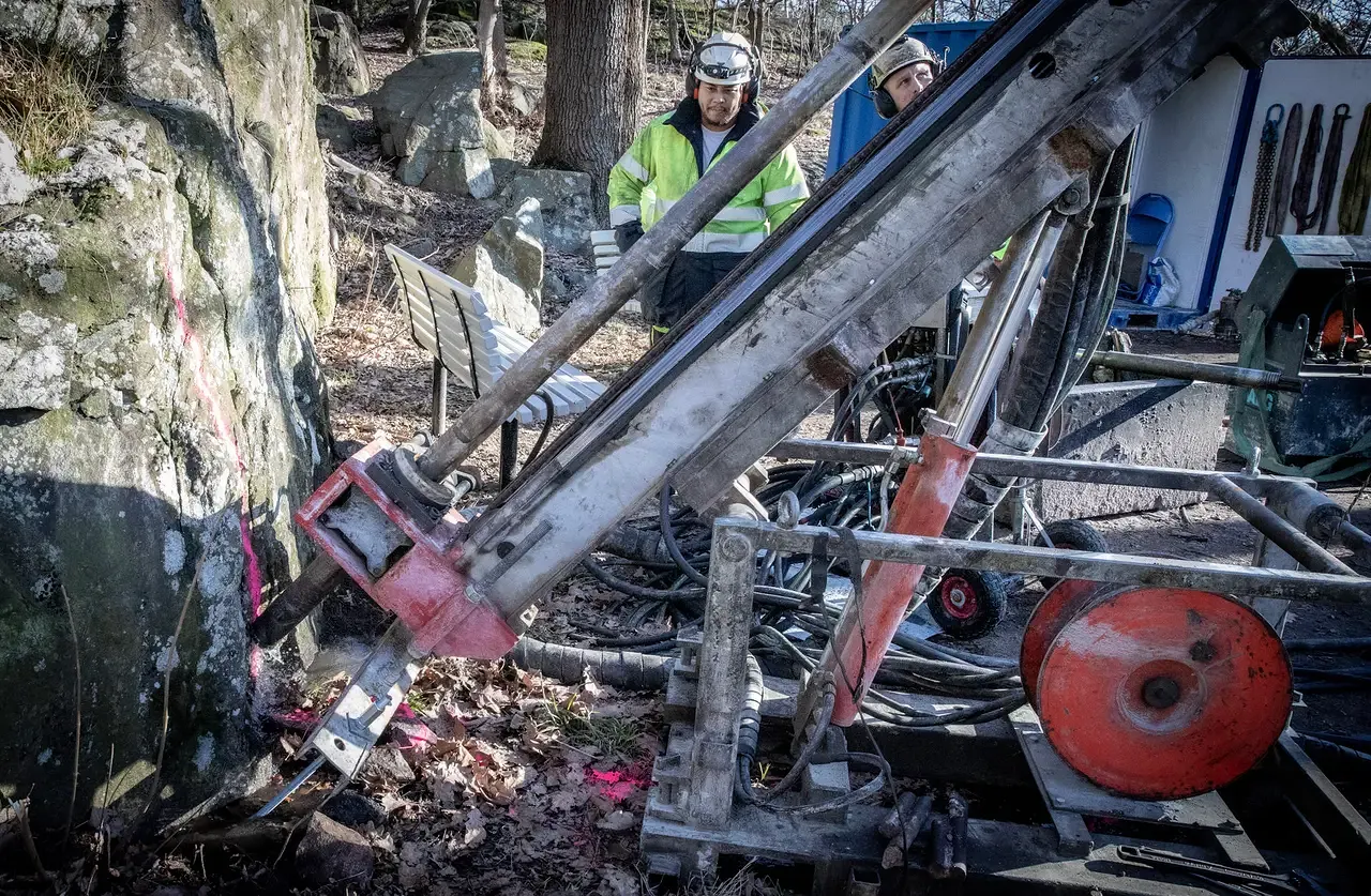 Två arbetare med hjälmar hanterar borrmaskin mot klippvägg i skog.